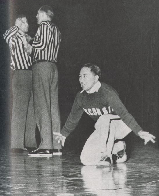Toru Iura performing during his time as a UW Cheerleader. He is kneeling on one knee in front of two referees. 